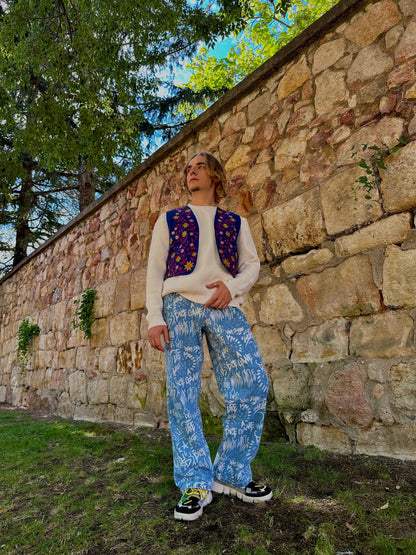 Person wearing a patterned sweater and pants standing against a stone wall with trees in the background