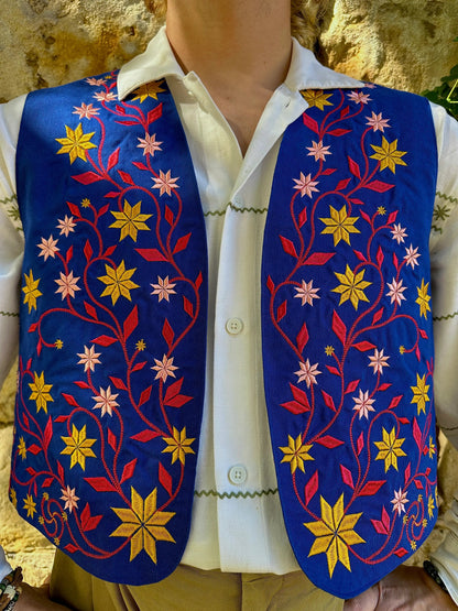 Mannequin wearing a white shirt with a colorful embroidered vest against a stone wall background
