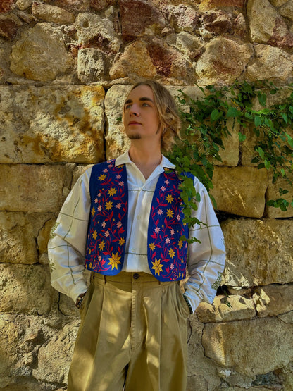 Person wearing a colorful vest and beige pants against a stone wall with greenery.