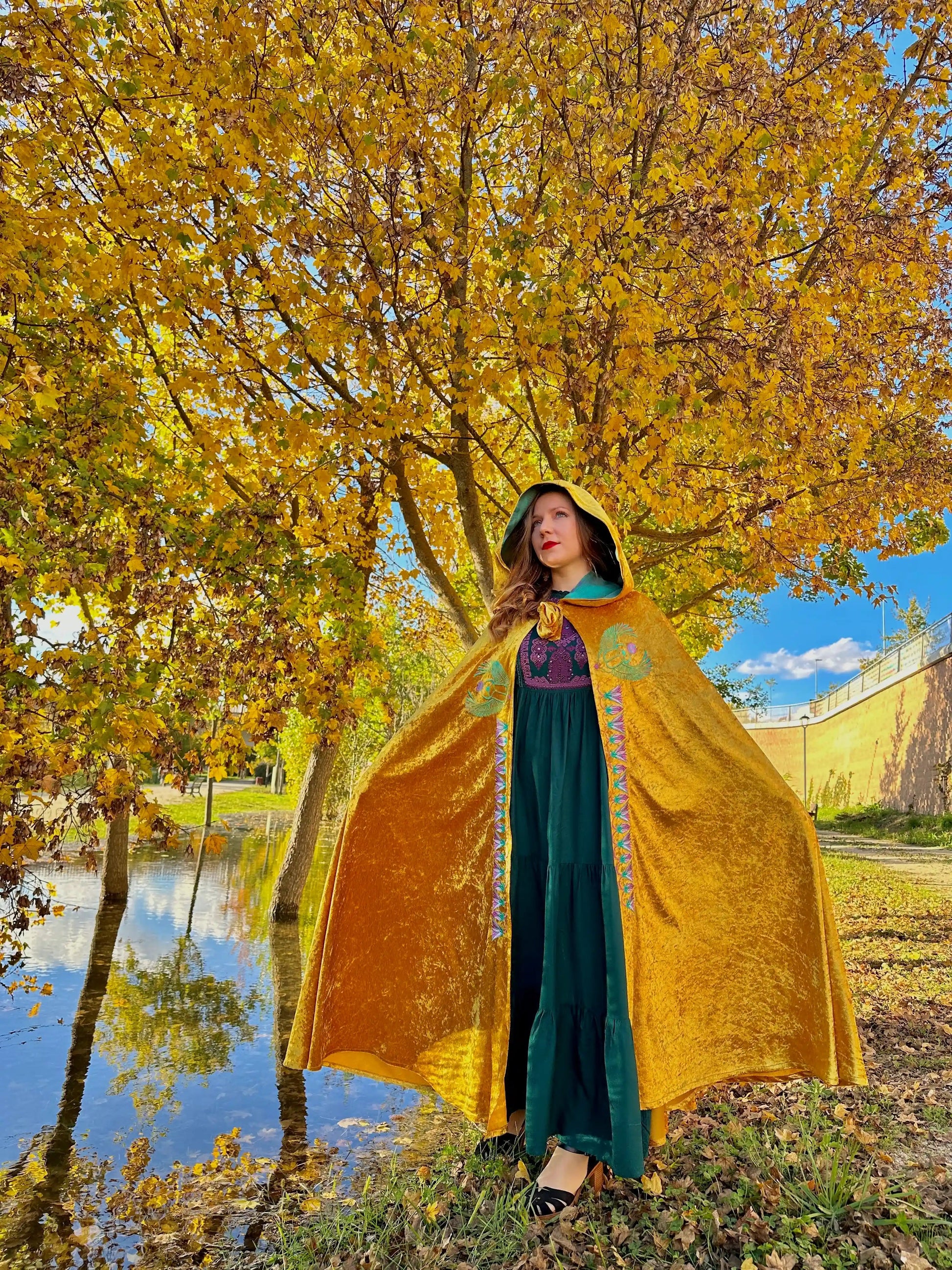 Person wearing a yellow cape and green dress standing in front of a tree with yellow leaves.