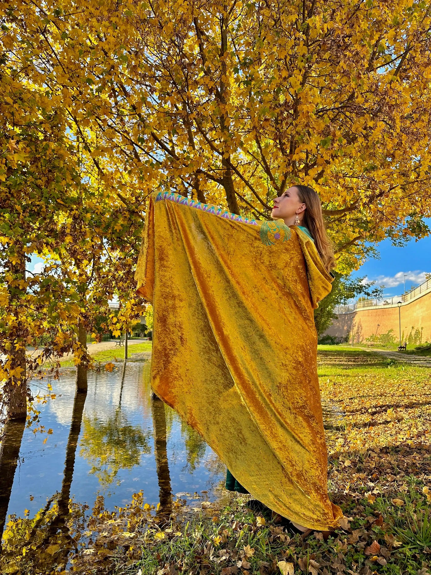 Woman holding a yellow fabric in an autumn park with trees and a pond.