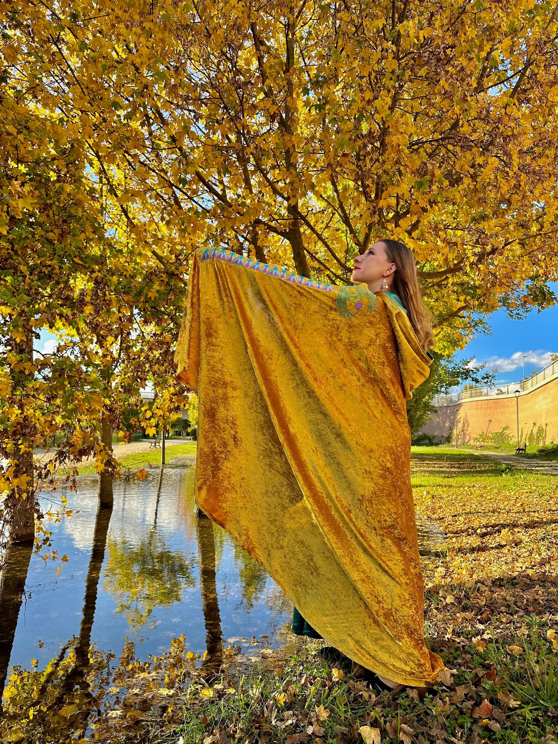 Woman holding a yellow fabric in an autumn park with trees and a pond.