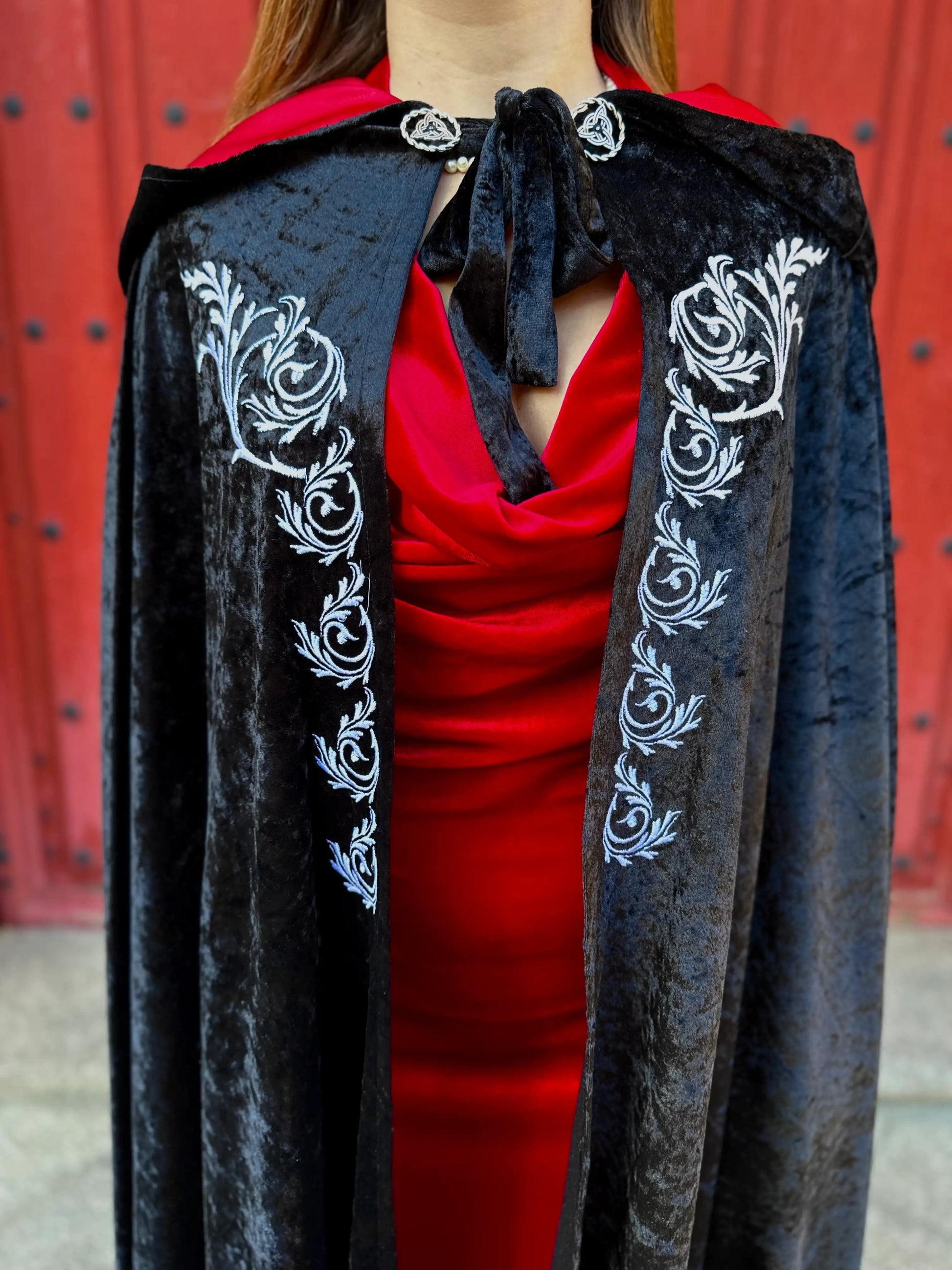 Black velvet cape with white floral embroidery worn over a red outfit against a red background