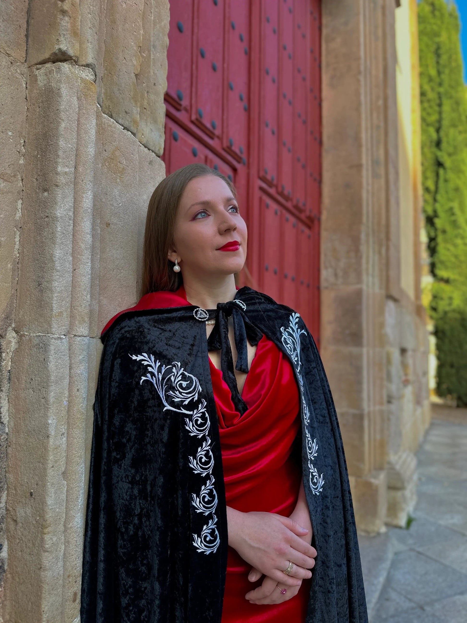 Woman in a red dress and black embroidered cape standing against a stone wall with a red door.