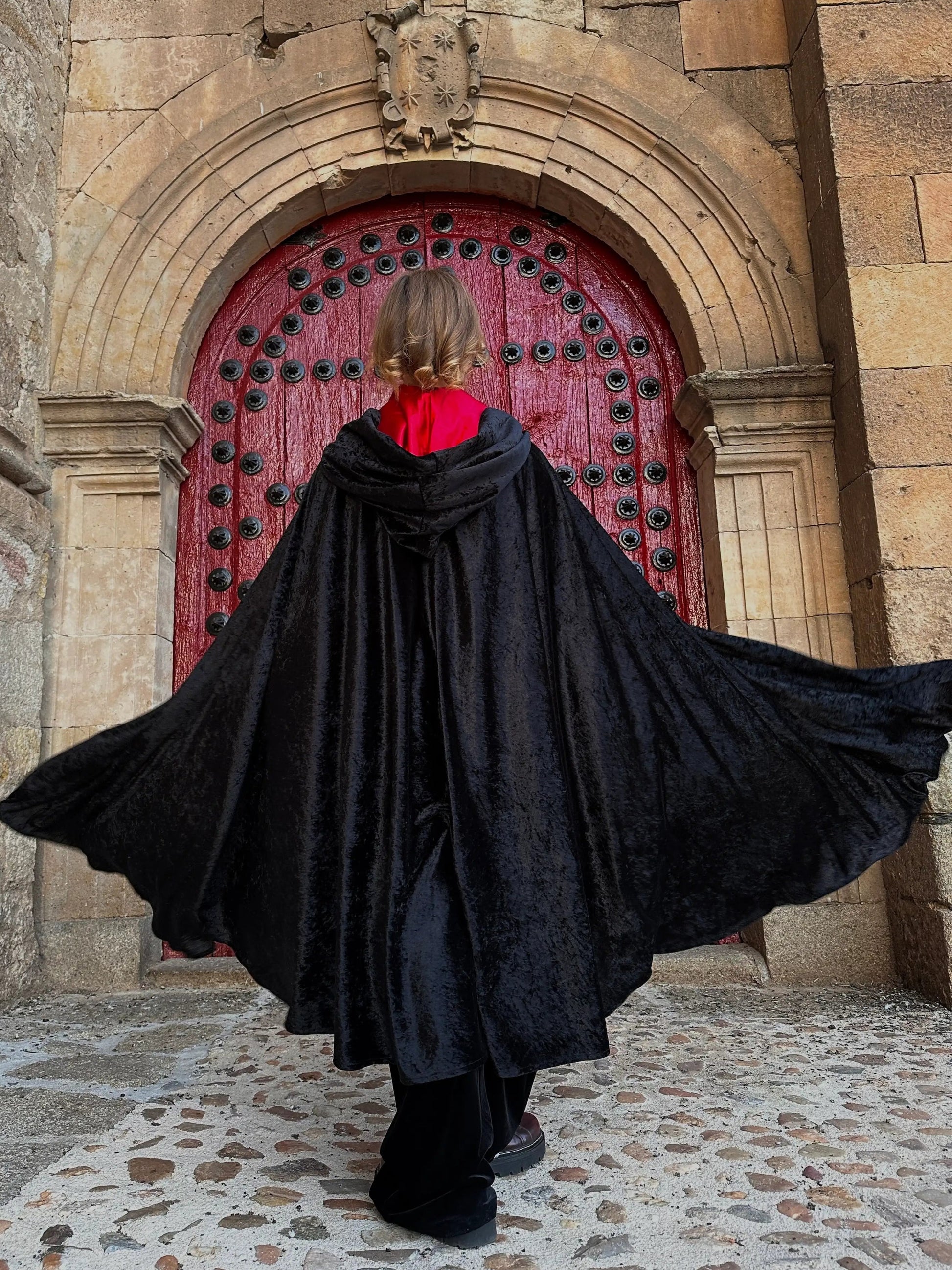 Person in a black cloak standing in front of a red door with stone architecture