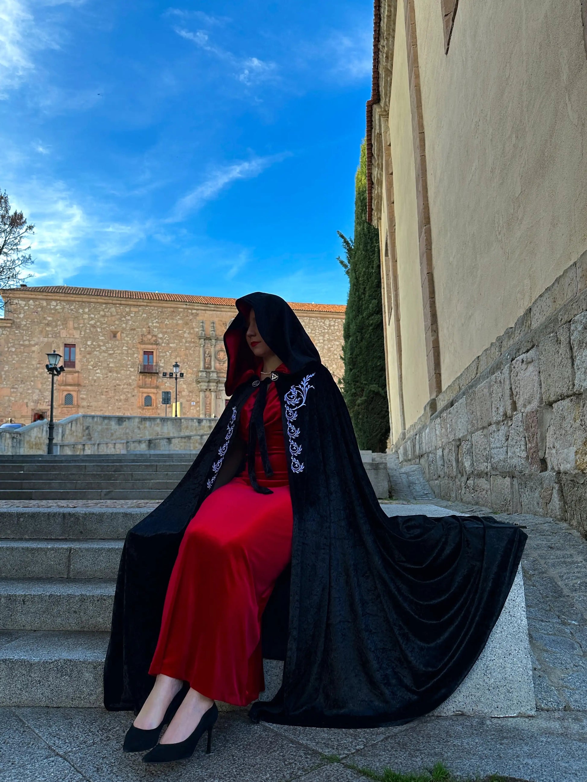 Woman in a red dress and black cape sitting on stone steps with a building and blue sky in the background