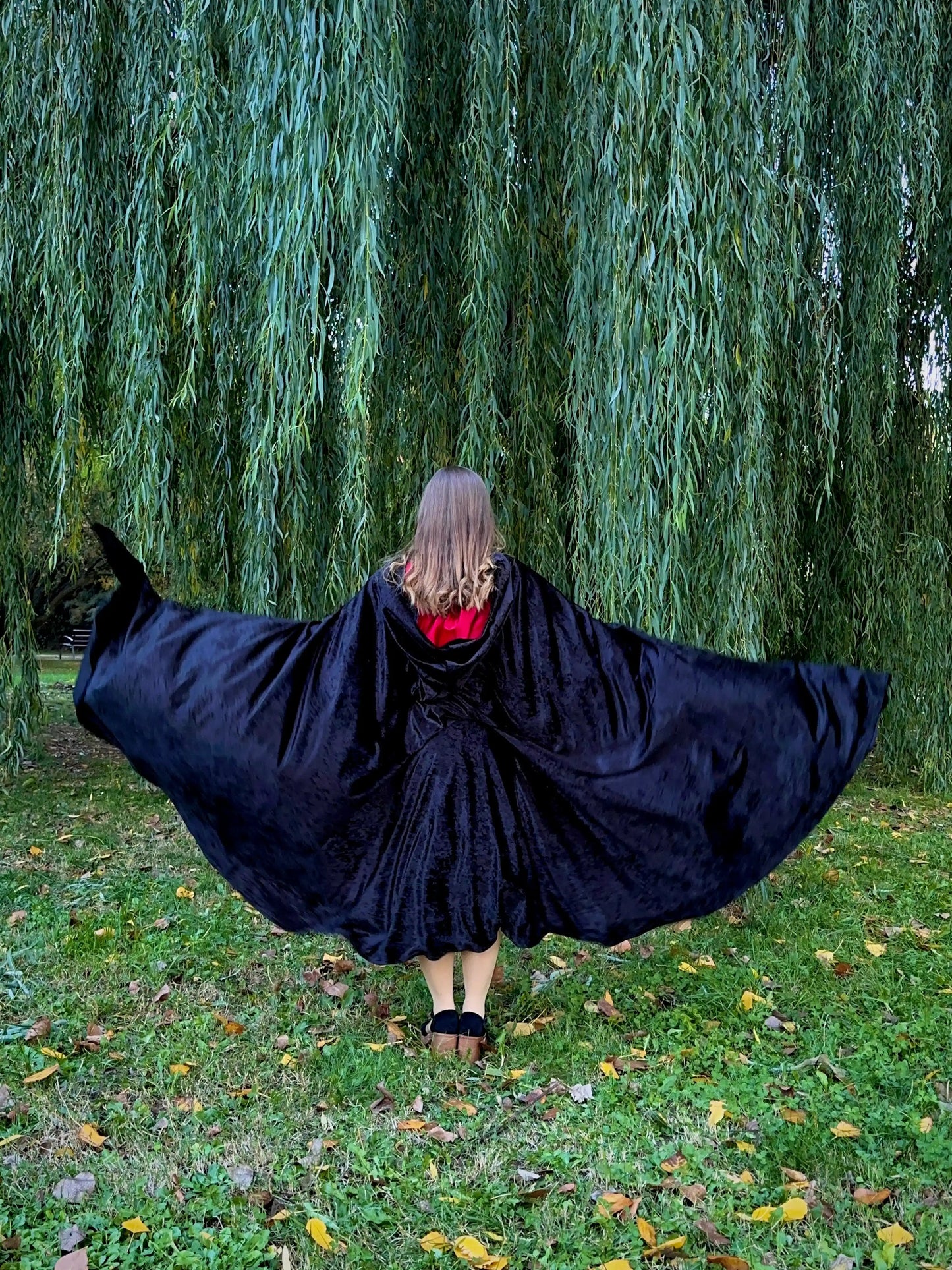Person wearing a black cape in a park with willow trees
