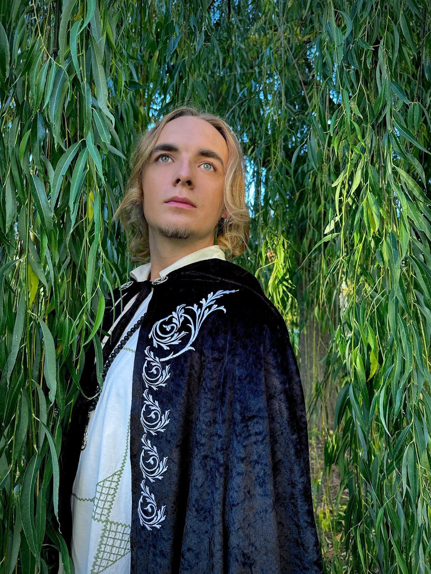 Person wearing a dark cloak with white embroidery standing among green trees