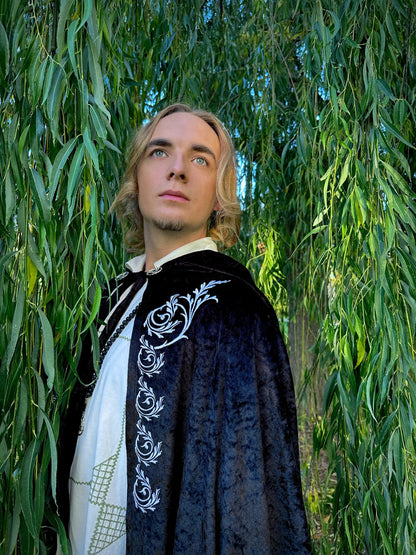 Person wearing a dark cloak with white embroidery standing among green trees