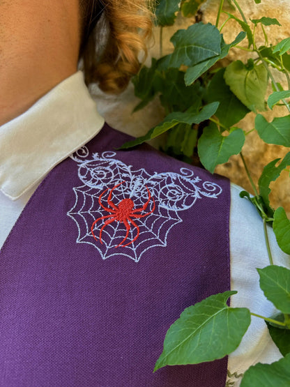 Purple garment with embroidered spider and web design, surrounded by green leaves.