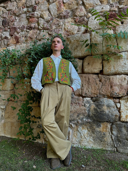 Man in traditional attire standing against a stone wall with greenery.