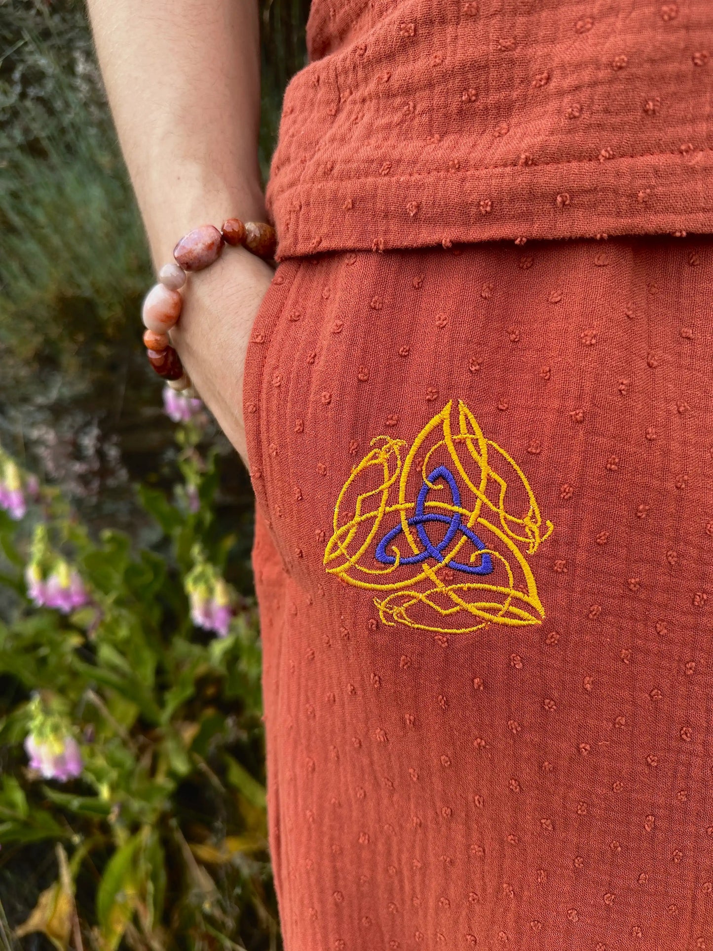 Person wearing a rust-colored garment with a Celtic knot design, holding a beaded bracelet against a natural background.