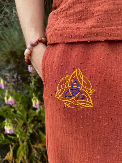 Person wearing a rust-colored garment with a Celtic knot design, holding a beaded bracelet against a natural background.