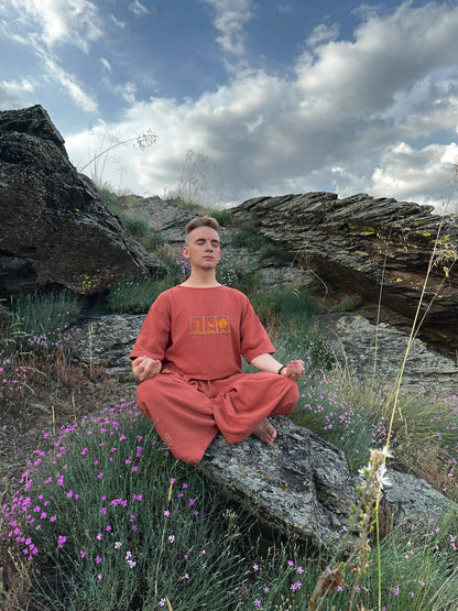 Person in orange outfit meditating on a rocky landscape with purple flowers and blue sky.