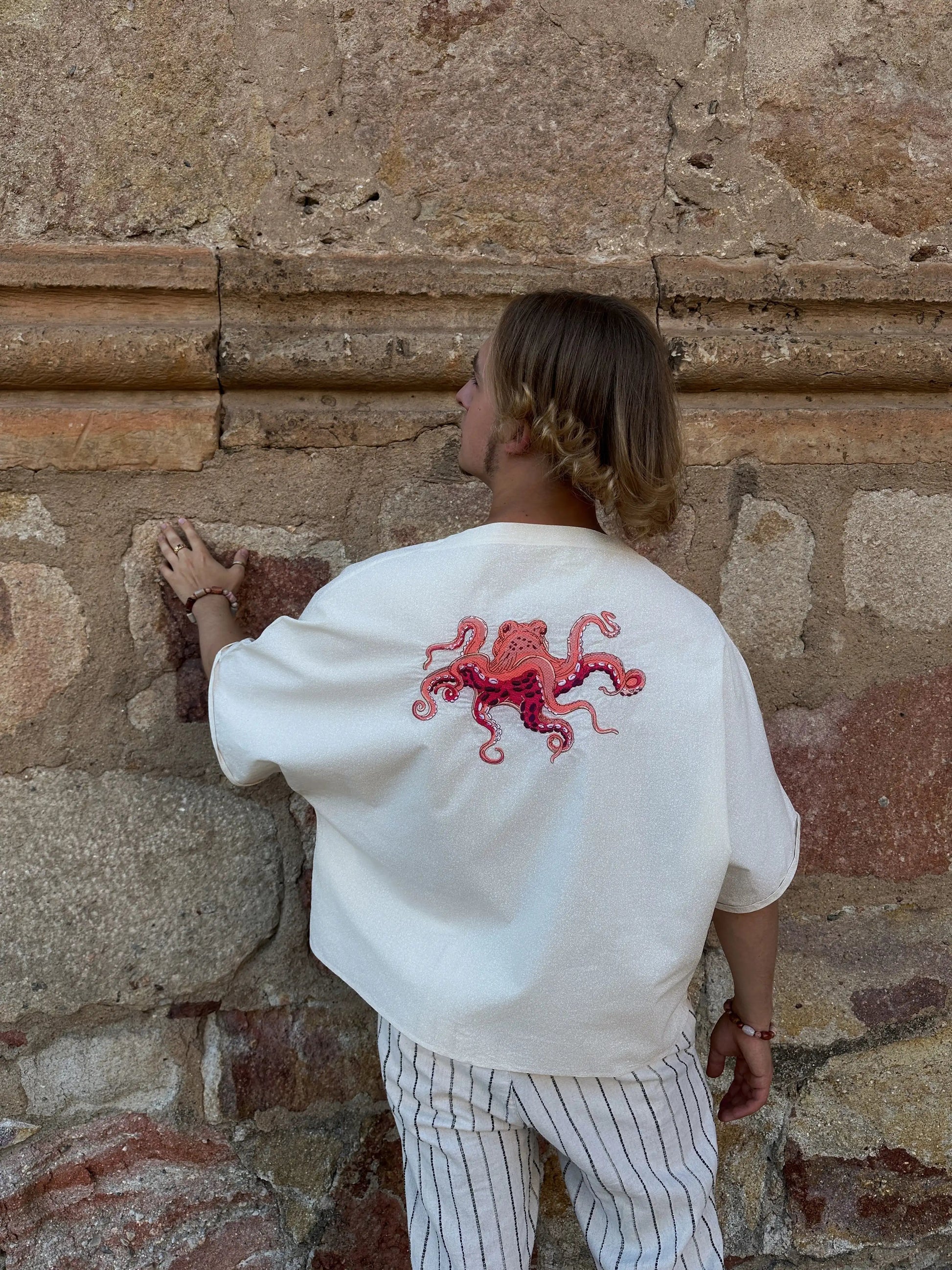 Person wearing a white sweatshirt with a red octopus design against a stone wall.
