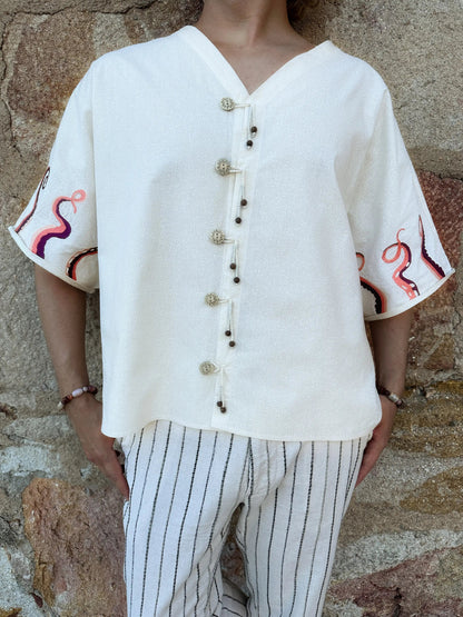 Person wearing a white blouse with embroidered sleeves against a stone wall.