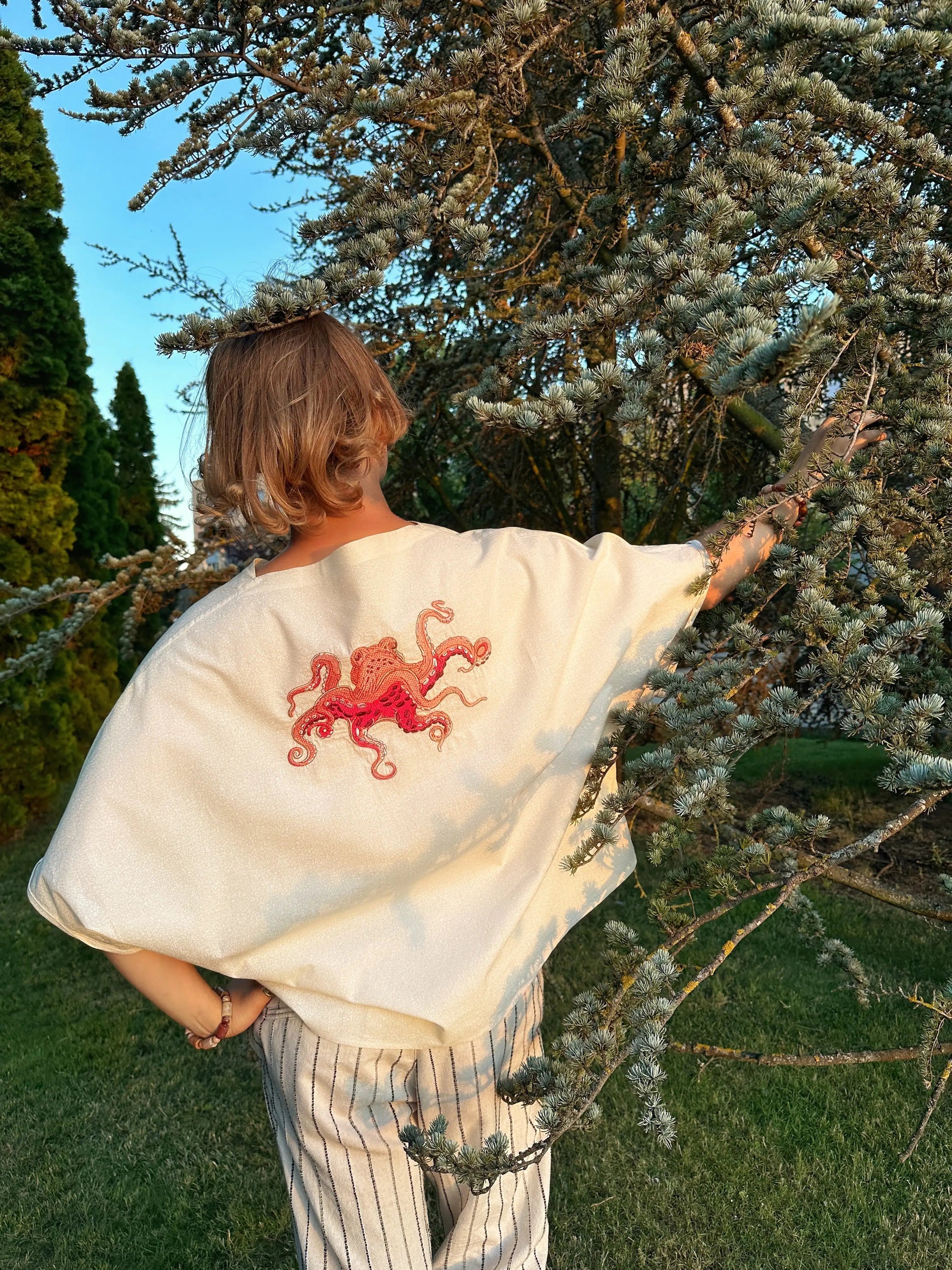 Person wearing a white shawl with a red embroidered design, standing outdoors near trees.