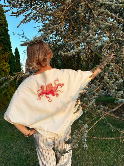 Person wearing a white shawl with a red embroidered design, standing outdoors near trees.