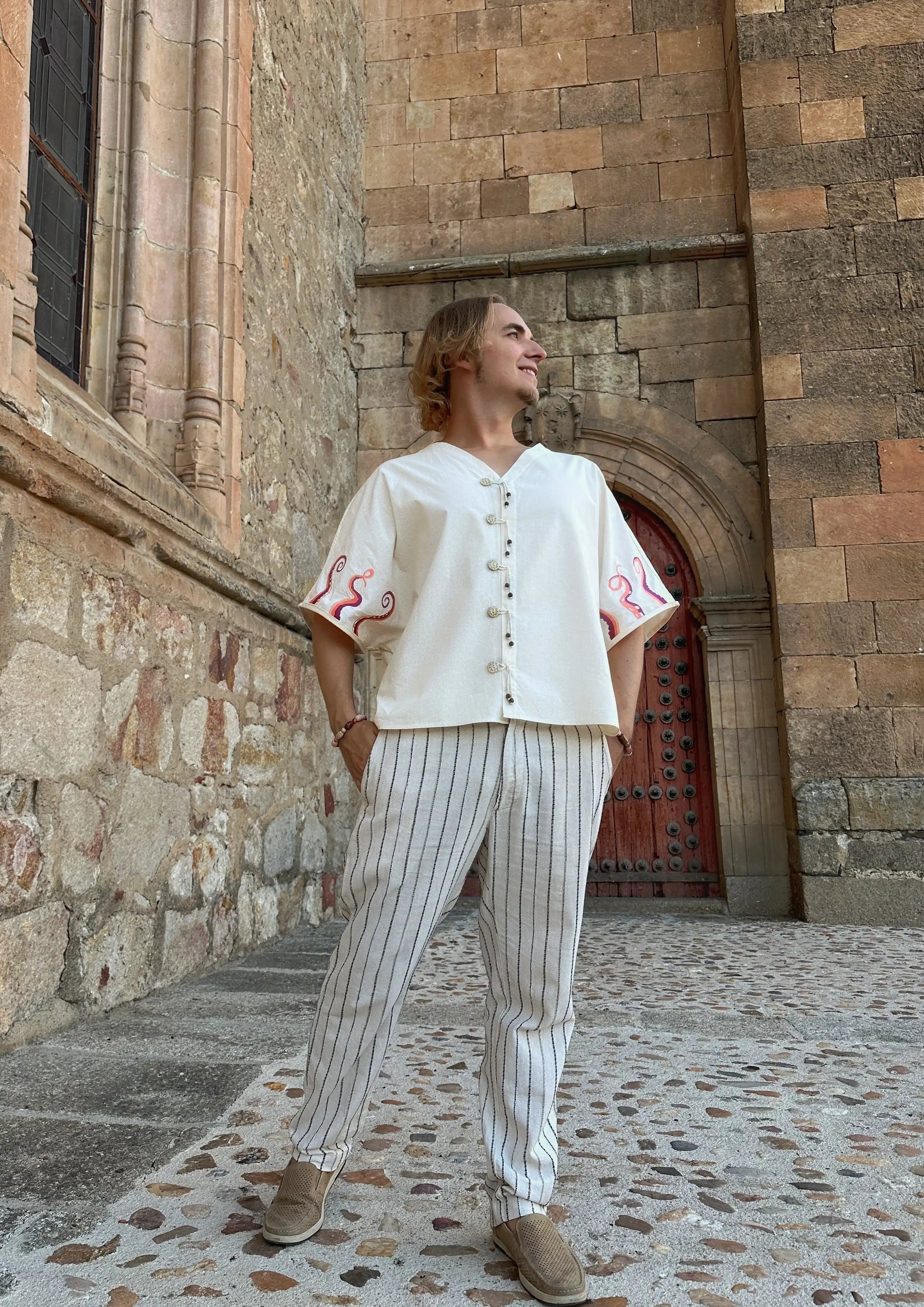 Person wearing a white outfit with red patterns standing in front of a stone building.