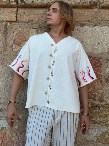 Person wearing a white embroidered shirt and striped pants standing against a stone wall.