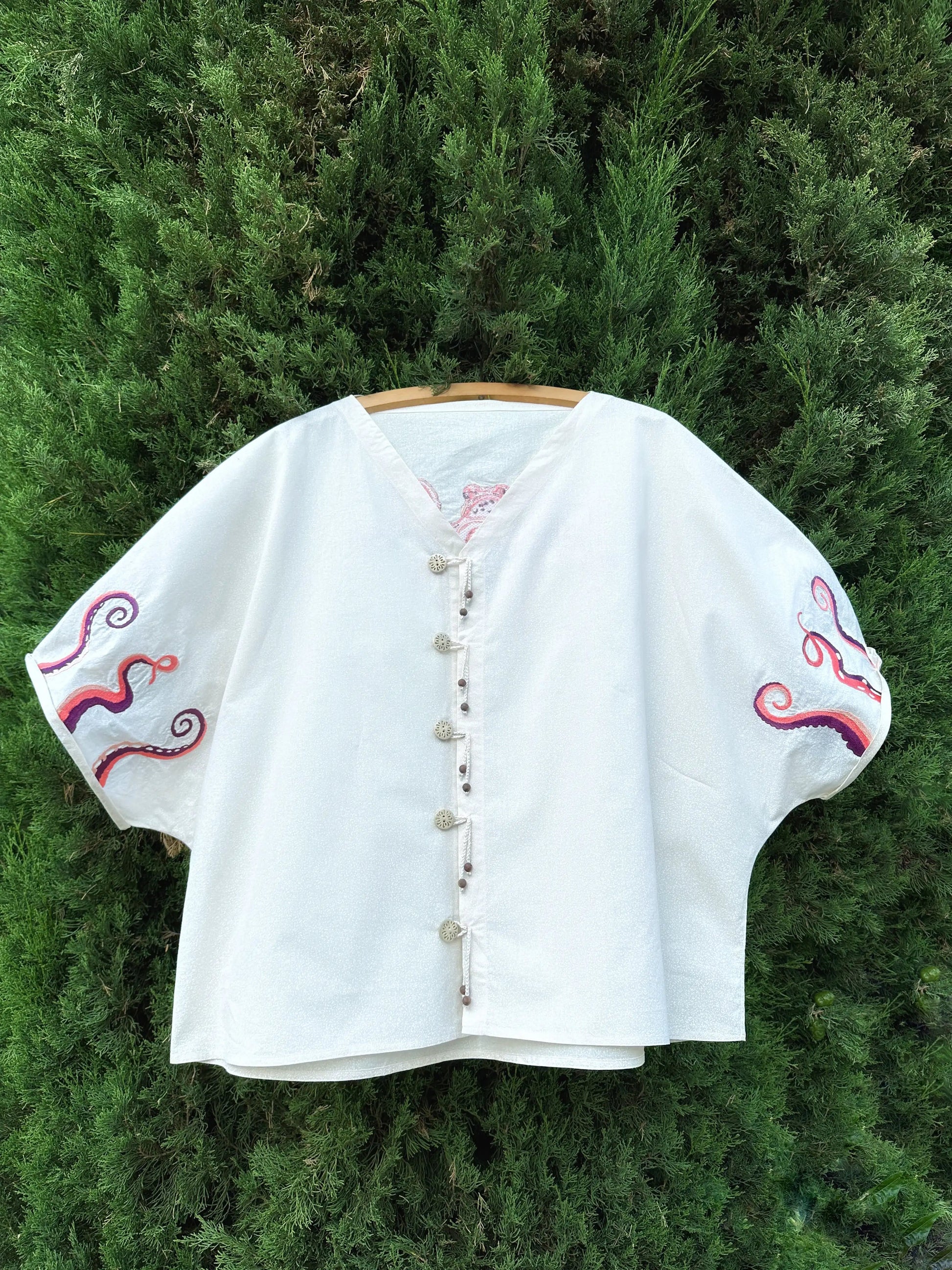 White embroidered blouse on a wooden hanger against a green bush background