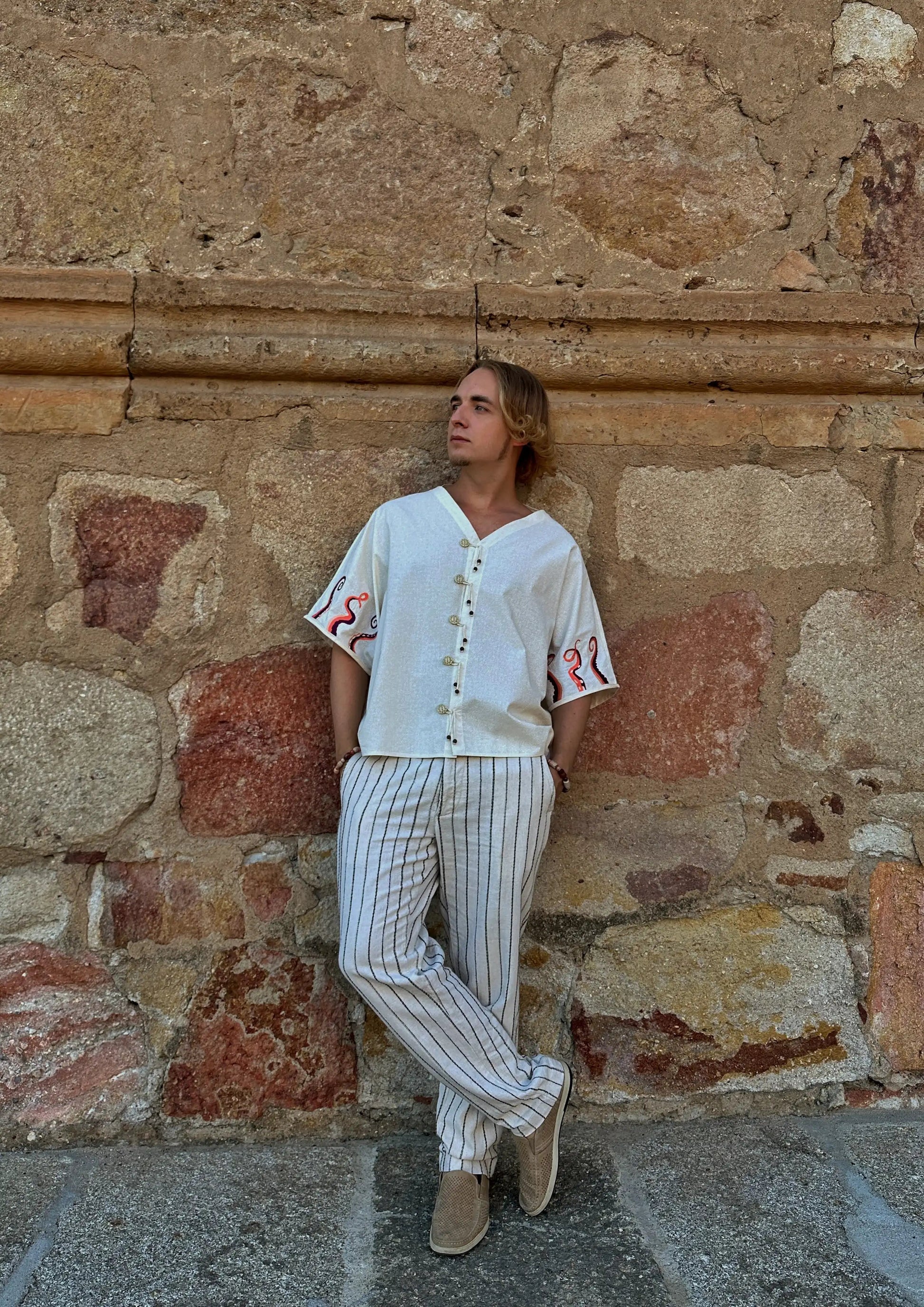 Person wearing a white shirt with red accents and striped pants against a stone wall.