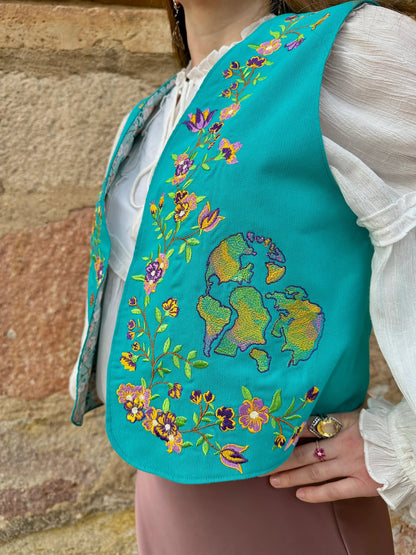 Teal embroidered vest with floral patterns worn by a person against a stone wall background