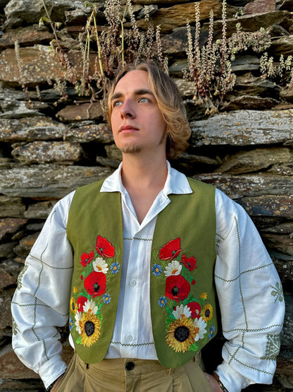 Man wearing a green embroidered vest with floral patterns against a stone wall.